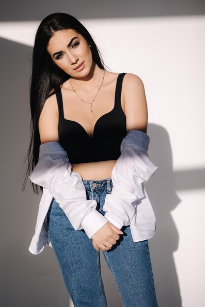 Pretty brunette hair woman in blue jeans and white shirt standing in front of white wall shadow on