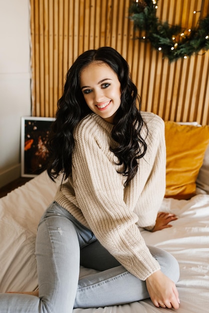 A pretty brunette in jeans and a beige sweater is sitting on the bed in the bedroom