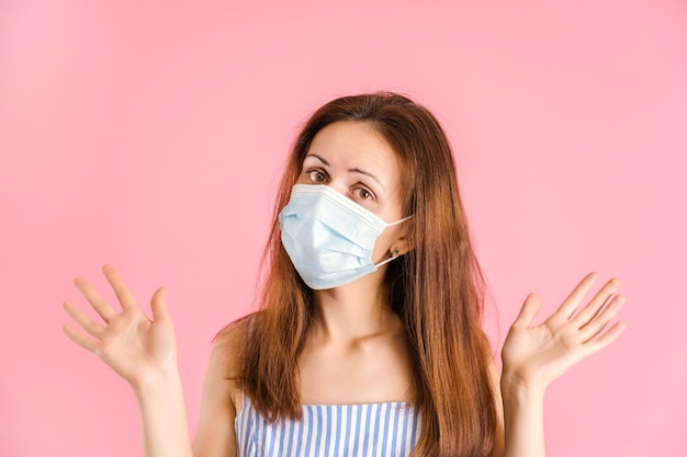 A pretty brunette in a medical mask gestures with her arms out to the sides. studio shot on a pink background