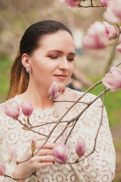 Pretty brunette model with nude makeup, wearing lace blouse, posing near the blooming magnolia flowers