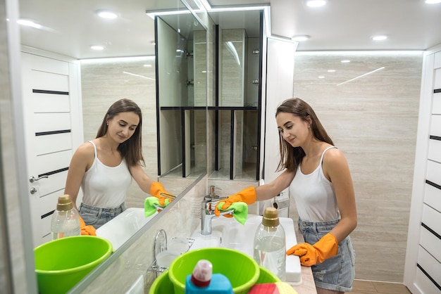 Pretty brunette in protective gloves cleans mirror in bath