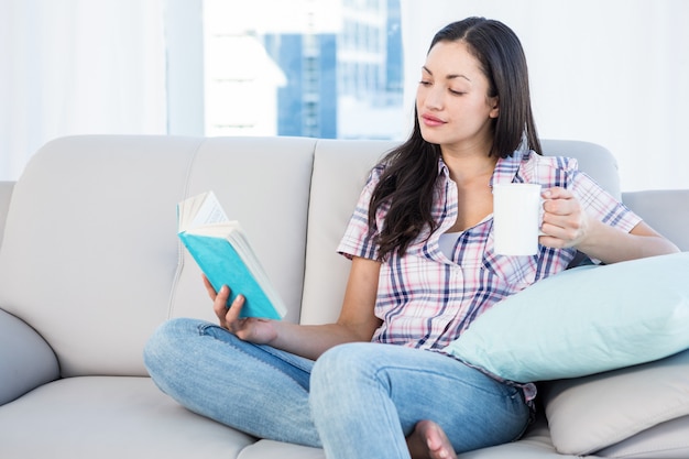 Pretty brunette reading and holding a cup of tea on couch in the living-room