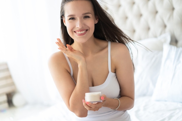 Pretty brunette using body cream on bed at home.