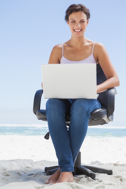 Pretty brunette using laptop on the beach sitting on swivel chair on a sunny day