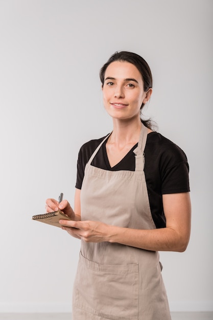 Pretty brunette waitress of cafe in apron making notes in small notepad while standing in front of camera in isolation