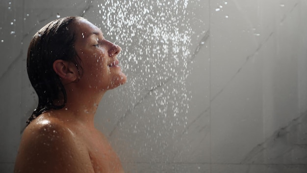 Фото Симпатичная брюнетка с голыми плечами и длинными мокрыми волосами стоит под струями душа за стеклянной копией пространства в ванной