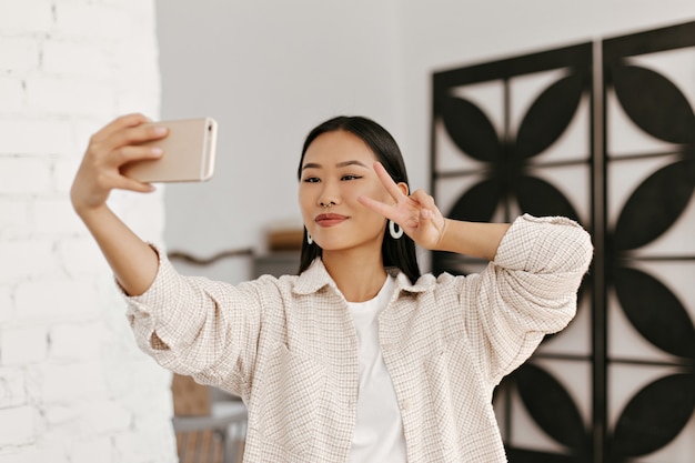 Pretty brunette woman in beige jacket and white t-shirt takes selfie and shows v-sign