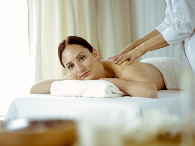 Pretty brunette woman enjoying procedure of back massage in spa salon. Beauty concept.