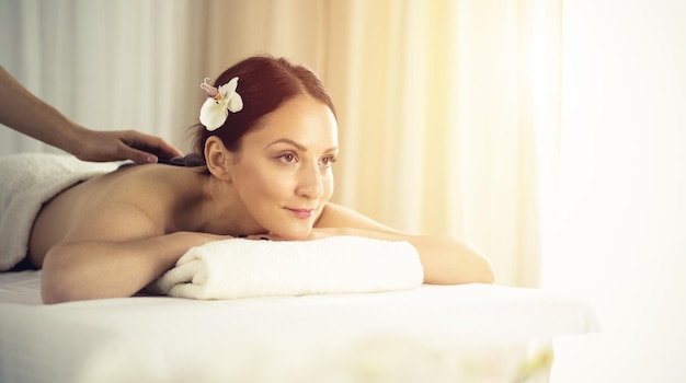 Pretty brunette woman enjoying treatment with hot stones in spa salon. Beauty concept.