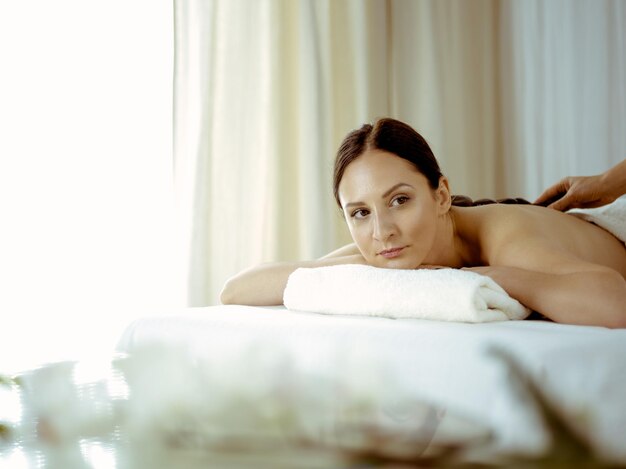 Pretty brunette woman enjoying treatment with hot stones in spa salon. Beauty concept.