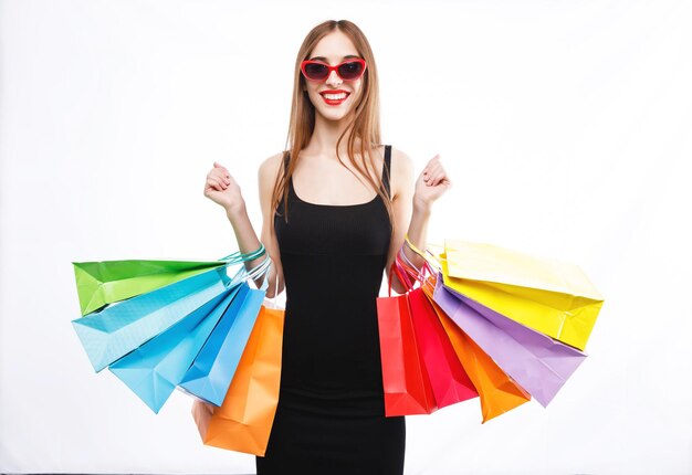 Pretty brunette woman wearing black dress and sunglasses holding colorful shopping bags and showing thumb up on the white background concept of consumerism sale rich life