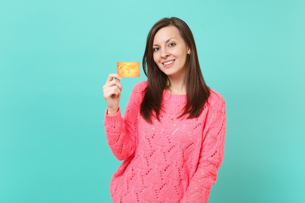 Pretty brunette young woman in knitted pink sweater looking camera holding in hand credit card isolated on blue turquoise wall background studio portrait. People lifestyle concept. Mock up copy space.