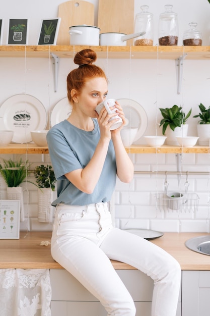 Pretty business lady drinking tasty beverage from mug at home office in during break Happy redhead young woman holding cup of hot coffee while standing in kitchen room with white modern interior