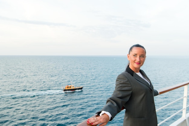 Pretty business woman or happy cute girl with smiling face in grey jacket, has red manicure on ship deck looking at sea, ocean water with sailing boat on cloudy sky background, traveling and tourism