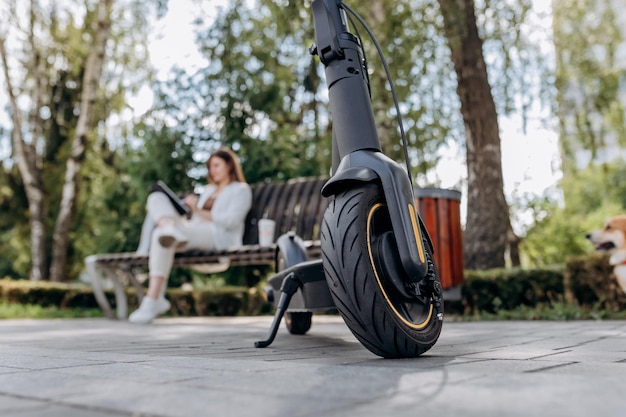 Photo pretty business woman in a white suit sits in a city park drinks coffee and works on a digital tablet with an electric scooter in the foreground