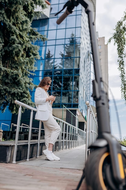 Bella donna d'affari in abito bianco e occhiali da sole che usa il telefono durante la pausa in piedi vicino a un moderno edificio per uffici con scooter elettrico in primo piano