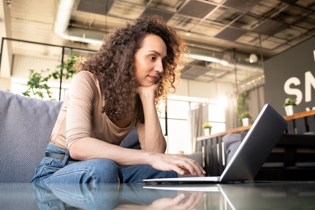 Pretty businesswoman in casualwear looking at laptop display while sitting on couch in cafe