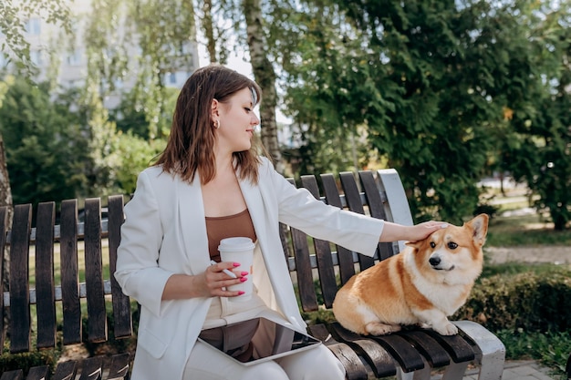 市内の公園に座ってコーヒーを飲み、犬のウェルシュ・コーギー・ペンブロークと一緒にデジタルタブレットに取り組んでいる白いスーツを着たかなりのビジネスウーマン