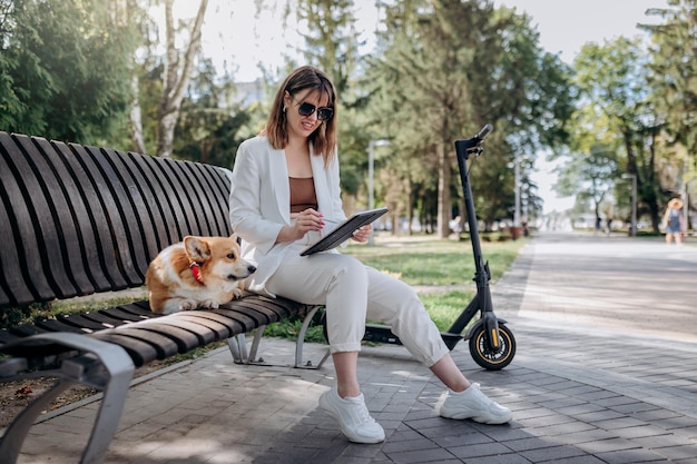 Pretty businesswoman in white suit sitting in city parkland with Welsh Corgi Pembroke dog and working on digital tablet with electric scooter on backgroundxA