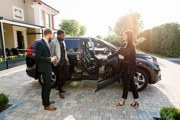 Pretty car saleswoman in business wear helping to young two multiethnical businessmen clients to make decision showing a new car, opening car doors, while standing on the yard of car salon outdoors