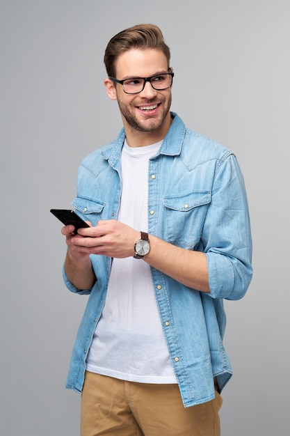 pretty casual man in blue jeans shirt holding his phone standing over studio grey wall