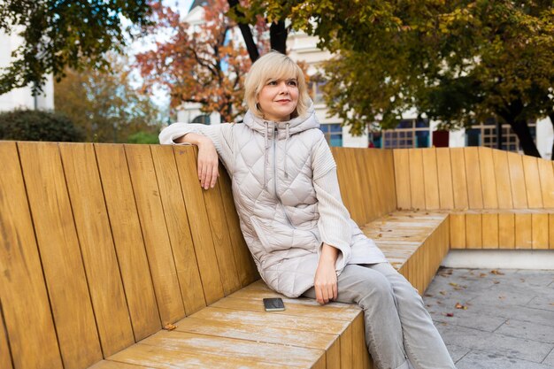 Pretty caucasian adult smiling blonde woman in jacket posing sitting on bench in city park concept o