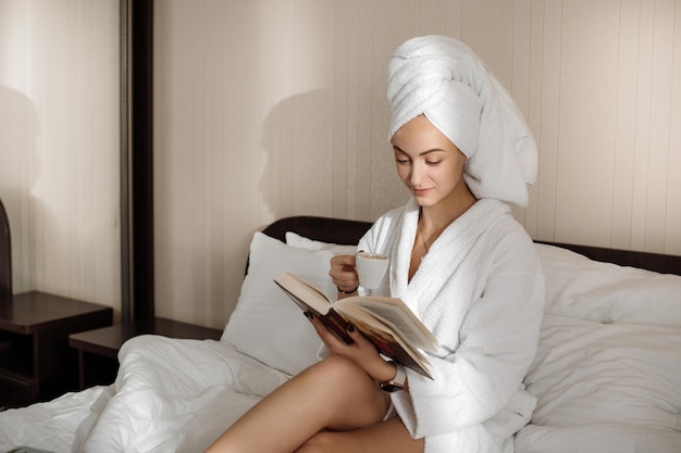 Pretty caucasian female in bathrobe and towel on head is reading a book on the bed