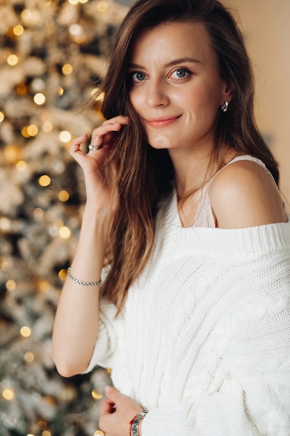 Pretty caucasian girl in white dress and stockings near the christmas tree with a lot of warm lights