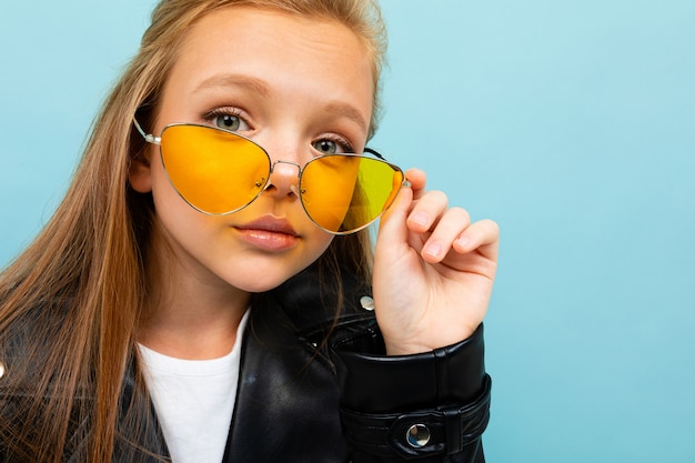 Pretty caucasian teenager girl with long brown hair in black jacket and denim jeans holds yellow sunglasses isolated on blue