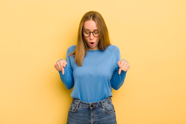 Pretty caucasian woman feeling shocked openmouthed and amazed looking and pointing downwards in disbelief and surprise