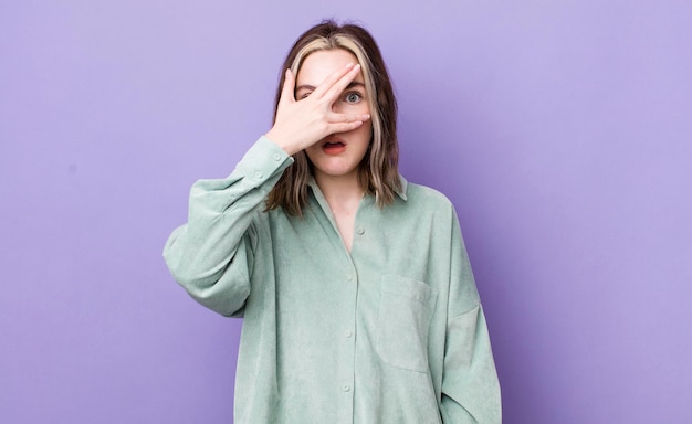 Pretty caucasian woman looking shocked scared or terrified covering face with hand and peeking between fingers