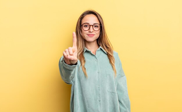 Photo pretty caucasian woman smiling and looking friendly, showing number one or first with hand forward, counting down