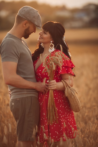 Pretty caucasian woman with long dark wavy hair in red dress with a bag and flowers hugs with her beautiful boyfriend in grey t-shirt, hat and shorts