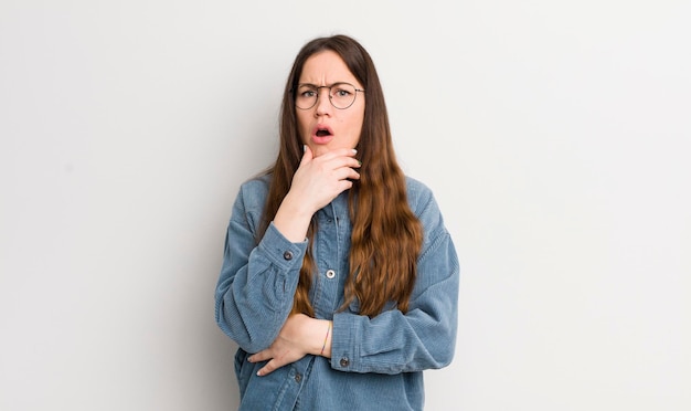 Pretty caucasian woman with mouth and eyes wide open and hand on chin feeling unpleasantly shocked saying what or wow