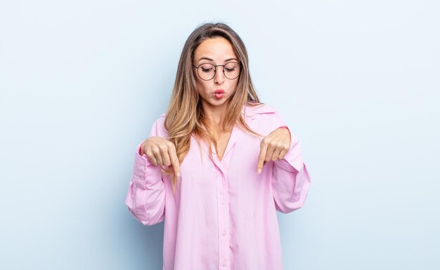 Pretty caucasian woman with open mouth pointing downwards with both hands, looking shocked, amazed and surprised