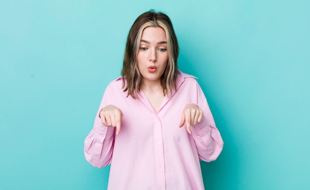 Pretty caucasian woman with open mouth pointing downwards with both hands looking shocked amazed and surprised