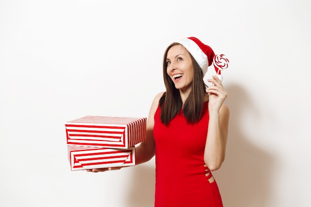 Pretty caucasian young happy woman with charming smile dressed in red dress and Christmas hat holding gift box and lollipopon white background. Santa girl with present isolated. New Year holiday 2018.