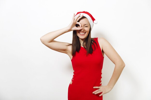 Pretty caucasian young happy woman with charming smile wearing red dress and Christmas hat showing OK gesture on white background. Santa girl making okay sign isolated. New Year holiday 2018 concept.