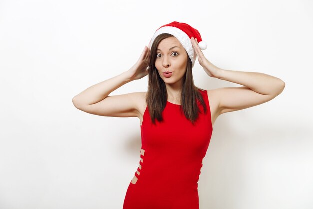 Giovane donna felice abbastanza caucasica con pelle sana e sorriso affascinante che indossa vestito rosso e cappello di natale che si tiene per mano sulla testa isolata su fondo bianco. ragazza di babbo natale. vacanze di capodanno 2018.