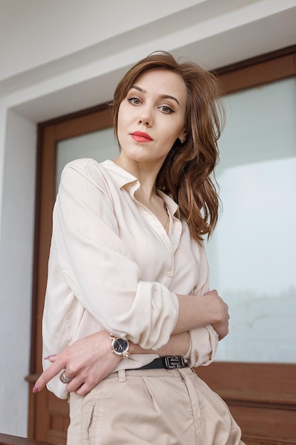 Pretty caucasian young woman in beige blouse, seductive red\
lipstick and wavy long hair standing outdoors near window of\
building, looking at camera