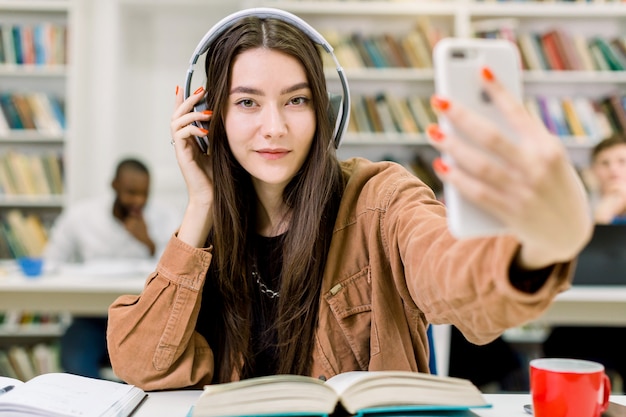 Pretty Caucasian young woman student in casual hipster clothes, making selfie photo on smartphone for sharing it in networks