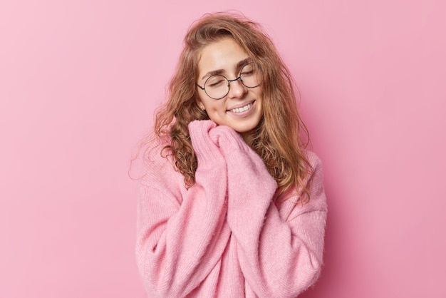 Pretty charming European woman smiles gently has dimples on cheeks keeps eyes closed keeps hands near face wears soft jumper isolated over pink background feels cozy and warm enjoys good mood