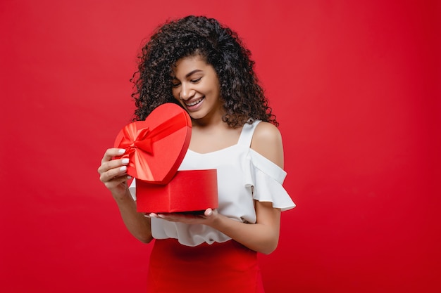 Pretty cheerful black woman opening heart shaped valentine gift isolated on red