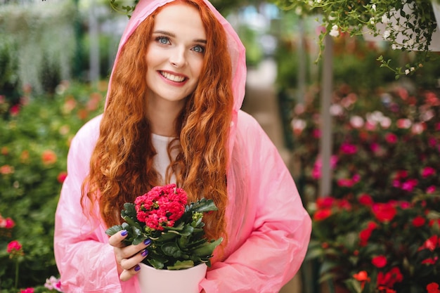 Pretty cheerful lady with redhead curly hair standing in pink raincoat and holding beautiful flower in pot while happily looking aside in greenhouse