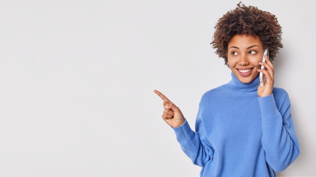 Pretty cheerful young woman makes telephone call holds smartphone near ear points away on blank copy space shows advertisement isolated over blue background. People technology promotion concept