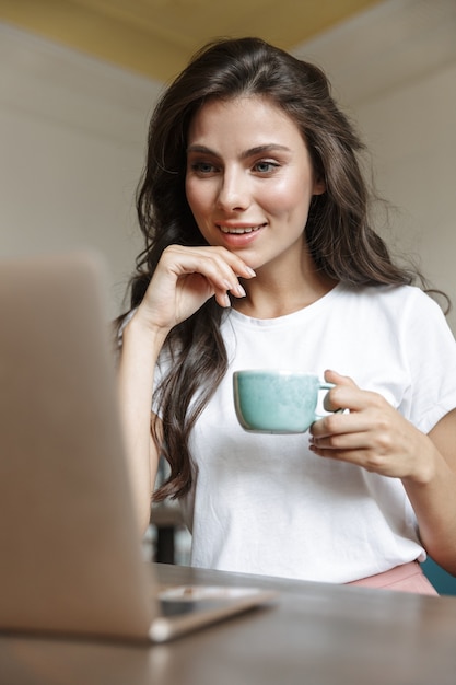 Довольно веселая счастливая молодая женщина в помещении дома с помощью портативного компьютера, пить кофе.
