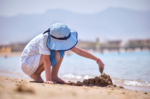 Pretty child girl in big hat and white dress playing with wet sand on background of blue sky and clear ocean lagoon water