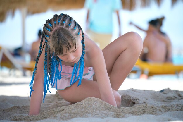 Ragazza graziosa del bambino in costume da bagno del bikini che gioca con la sabbia nell'ombra dell'ombrello durante le vacanze tropicali estive