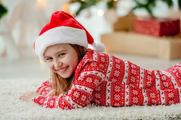 Милая детская девочка, лежащая на полу в доме Рождества в традиционной шляпе Сантаа. Счастливый улыбающийся ребенок в новогоднее время