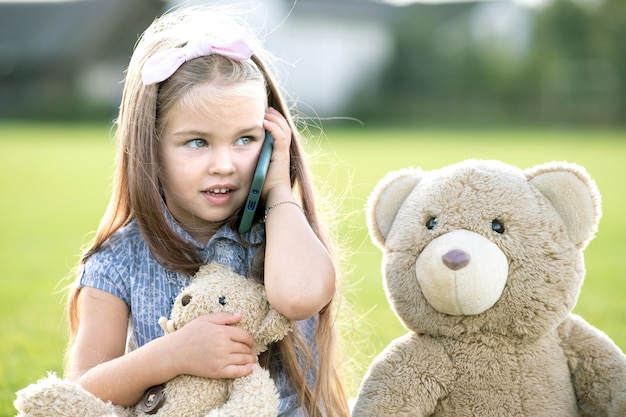 Pretty child girl sitting in summer park on green grass with her teddy bear toy talking on mobile phone outdoors in summer